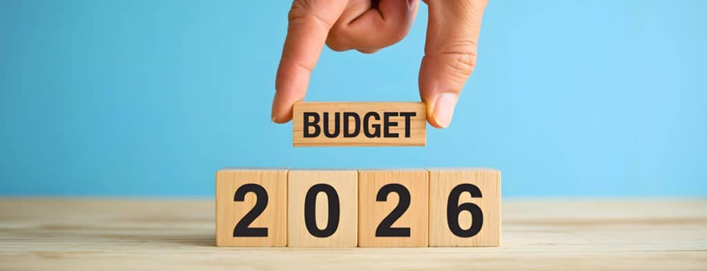 Hand placing a 'BUDGET' block above wooden blocks that spell '2026' against a blue background.