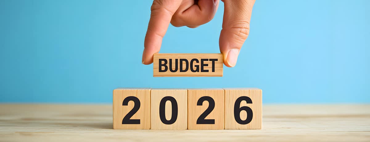 Hand placing a 'BUDGET' block above wooden blocks that spell '2026' against a blue background.
